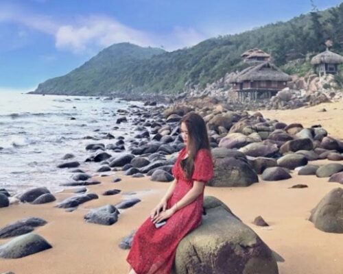 Cô gái trong chiếc váy đỏ ngồi yên bình trên tảng đá bên bãi biển, hướng ra biển.